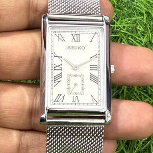 Puede incluir: Un reloj Seiko rectangular con una correa de malla plateada. La esfera es blanca con números romanos y agujas negras. Un dial más pequeño muestra los segundos. El reloj se sostiene sobre un fondo verde.