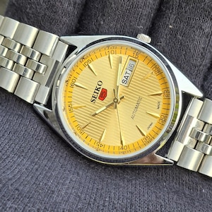 May include: A Seiko 5 automatic wristwatch with a gold-toned dial featuring a horizontal striped pattern, silver-toned hands and hour markers, and a day-date window at the three o'clock position. The watch has a stainless steel link bracelet.