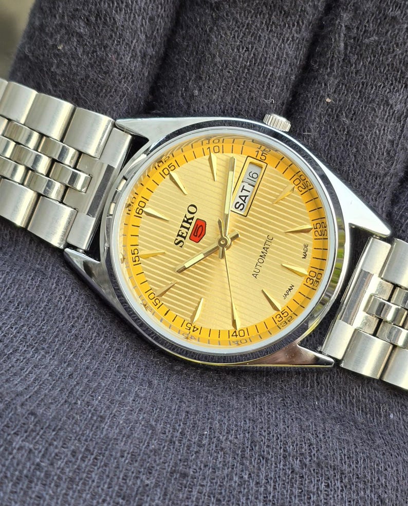 May include: A Seiko 5 automatic wristwatch with a gold-toned dial featuring a horizontal striped pattern, silver-toned hands and hour markers, and a day-date window at the three o'clock position. The watch has a stainless steel link bracelet.