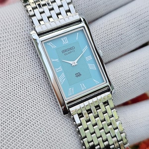 May include: A rectangular Seiko Quartz watch with a light blue dial featuring a wavy pattern, silver-tone Roman numeral hour markers, and silver hands. The watch has a polished silver-tone metal link bracelet with a multi-row design.