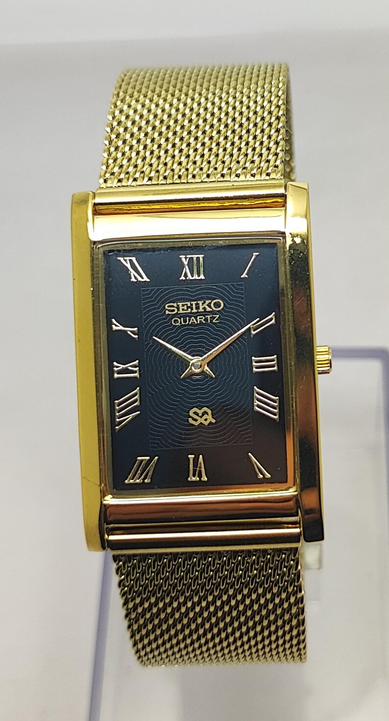 Peut inclure: Montre Seiko Quartz dor&eacute;e avec un cadran noir rectangulaire et des chiffres romains dor&eacute;s. La montre est dot&eacute;e d'un bracelet en maille et d'un bo&icirc;tier dor&eacute;. Le cadran affiche le logo Seiko Quartz et l'heure.