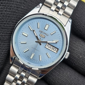May include: A Seiko 5 automatic wristwatch featuring a light blue sunray dial with silver-tone baton indices, luminous hands, and a day-date window at the three o'clock position. The timepiece includes a stainless steel case and a link bracelet.