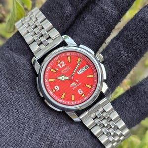 Pode incluir: Um relógio Citizen Automatic com mostrador vermelho, caixa prateada e pulseira de aço inoxidável. O mostrador mostra o dia da semana e a hora com ponteiros e marcadores luminosos. O relógio está sobre uma superfície cinza escuro.