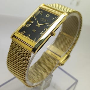 Peut inclure: Montre Seiko Quartz dor&eacute;e avec un cadran noir rectangulaire et des chiffres romains dor&eacute;s. Le bracelet est en maille et le bo&icirc;tier est dor&eacute;. La montre est pr&eacute;sent&eacute;e en biais.
