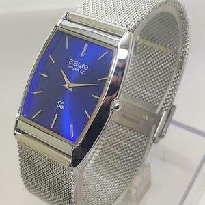 Może przedstawiać: Prostokątny zegarek Seiko Quartz z żywą niebieską tarczą i srebrnymi wskazówkami. Zegarek posiada bransoletę z siatki ze stali nierdzewnej i srebrną kopertę. Zegarek jest prezentowany na przezroczystym stojaku.