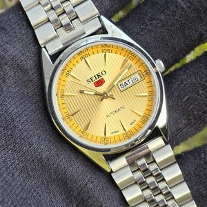 May include: A Seiko 5 automatic wristwatch with a gold-toned dial featuring a textured vertical stripe pattern, silver-toned hands and hour markers, and a day-date window at the three o'clock position. The watch has a silver-toned metal link bracelet.
