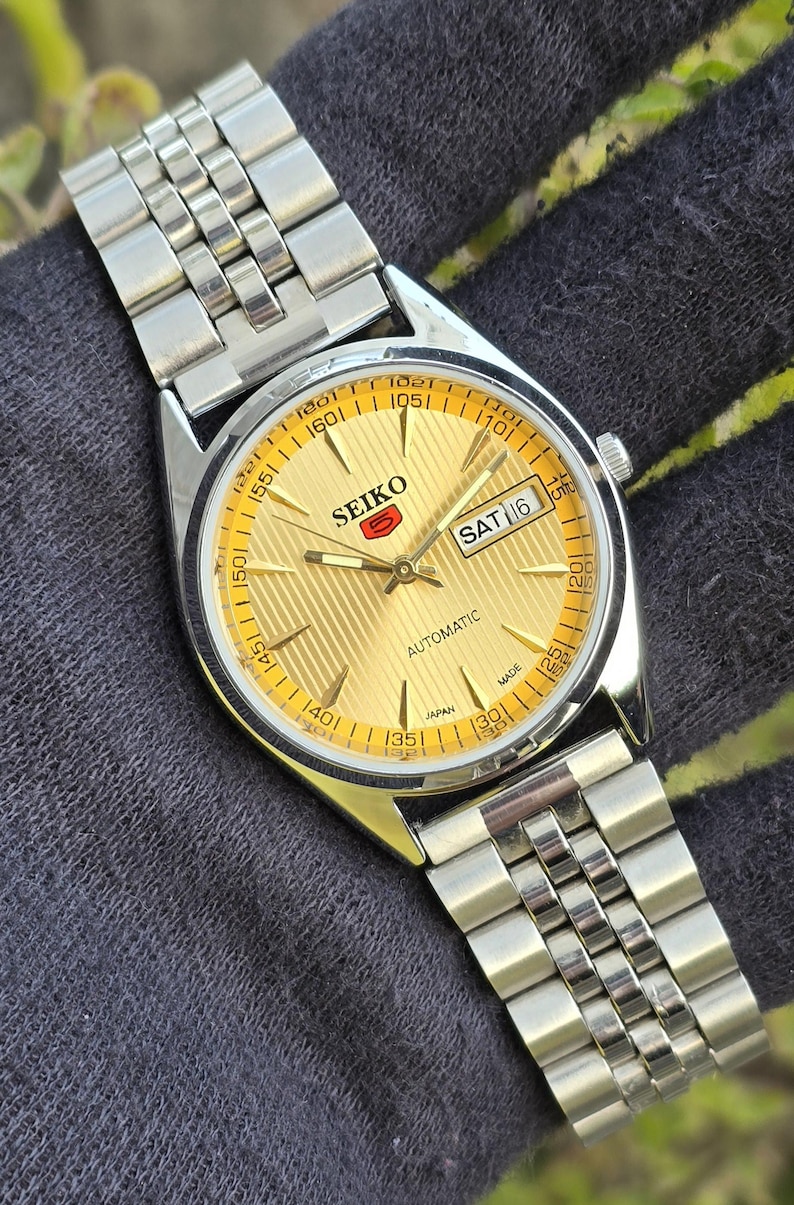 May include: A Seiko 5 automatic wristwatch with a gold-toned dial featuring a textured vertical stripe pattern, silver-toned hands and hour markers, and a day-date window at the three o'clock position. The watch has a silver-toned metal link bracelet.