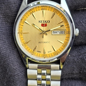May include: A Seiko 5 automatic wristwatch with a gold-toned textured dial, silver-toned hands, and hour markers. The watch features a day-date window at the three o'clock position, a silver-toned stainless steel link bracelet, and a tachymeter scale.