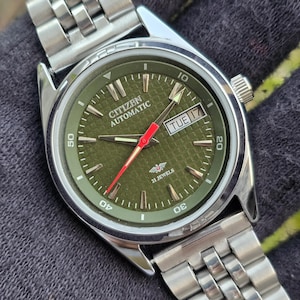 May include: A silver Citizen Automatic wristwatch with a green face and a silver strap. The watch shows the day and date, with a red second hand. The watch face has the text "Citizen Automatic" and "21 Jewels".