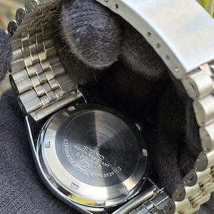 May include: A silver-tone stainless steel wristwatch with a metal link bracelet. The circular case back features engraved text including SEIKO, WATER RESISTANT, STAINLESS STEEL, and the model number 6309-5820, along with a serial number 335718.