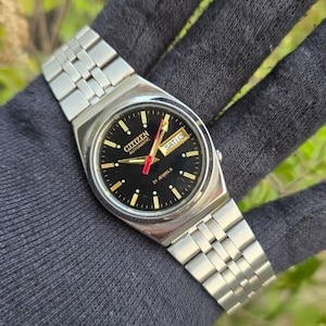 Pode incluir: Um relógio de pulso Citizen Automatic com pulseira de aço inoxidável prateada e mostrador preto. O mostrador apresenta marcadores de hora, ponteiros e visor de dia e data dourados. O relógio diz "Citizen Automatic 21 Jewels".