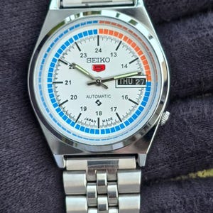 May include: A silver Seiko automatic wristwatch with a white face, featuring blue and orange accents. The watch displays the day and date, with a stainless steel band. The watch face includes the text "SEIKO" and "AUTOMATIC".