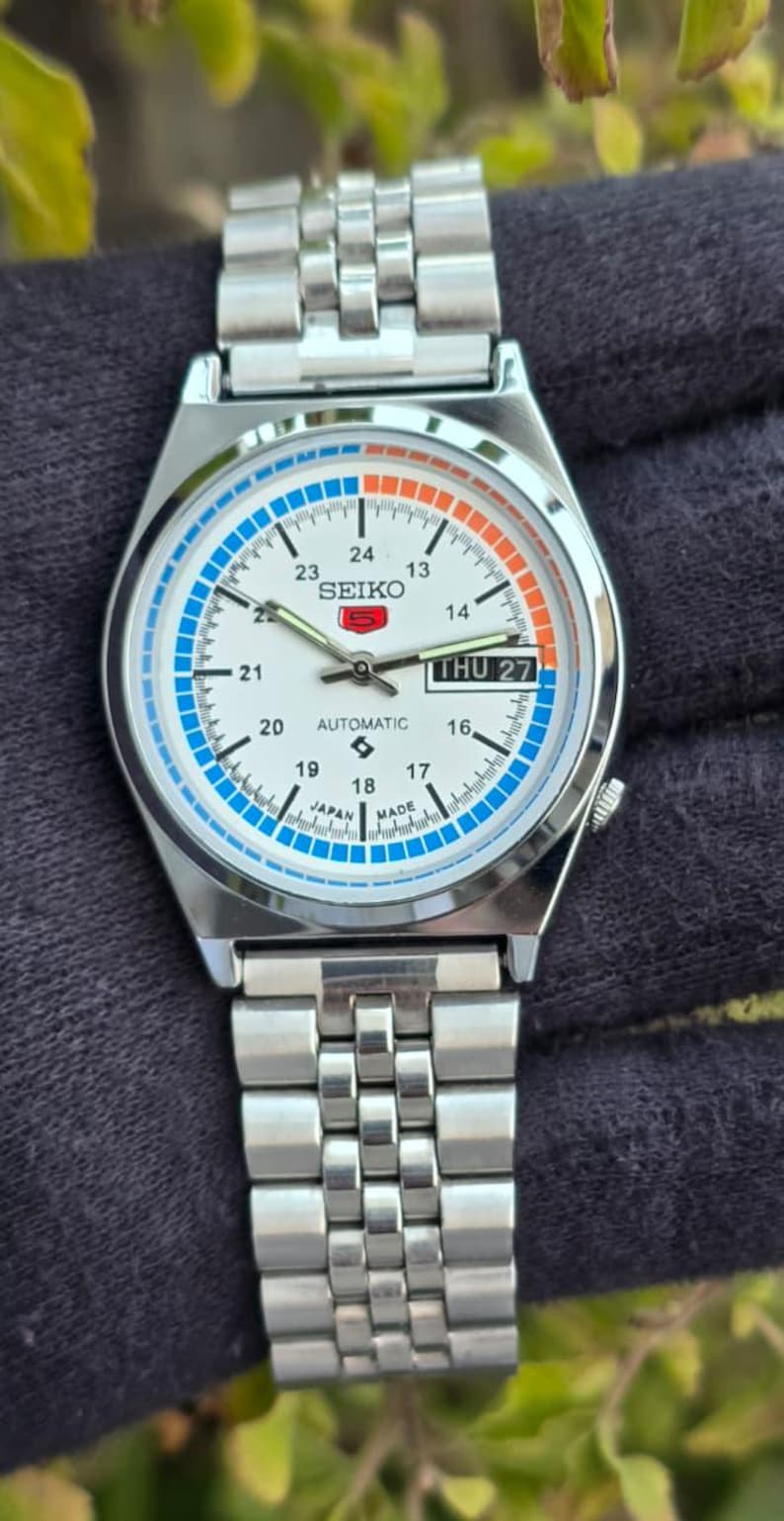 May include: A silver Seiko automatic wristwatch with a white face, featuring blue and orange accents. The watch displays the day and date, with a stainless steel band. The watch face includes the text "SEIKO" and "AUTOMATIC".