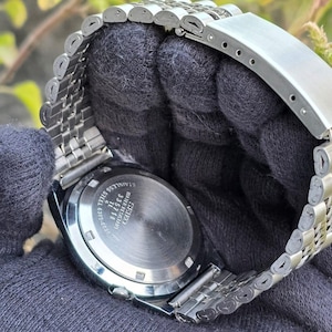 May include: A stainless steel Seiko wristwatch with a silver band. The watch face is not visible, but the back of the watch is visible, with the text "SEIKO" and other details. The watch is on a dark gray surface.