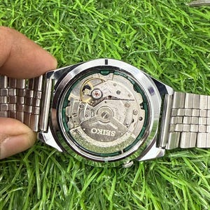 May include: A silver-toned Seiko watch with a transparent back, showcasing the intricate inner workings of the mechanism. The watch has a silver band and is set against a backdrop of green grass.