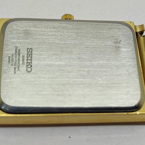 Peut inclure: Une montre Seiko dor&eacute;e avec un cadran rectangulaire et un bracelet en maille. Le bo&icirc;tier de la montre est dor&eacute;, et le dos est argent&eacute; avec le logo et le texte Seiko. Le bracelet en maille est &eacute;galement dor&eacute; et semble &ecirc;tre en m&eacute;tal.