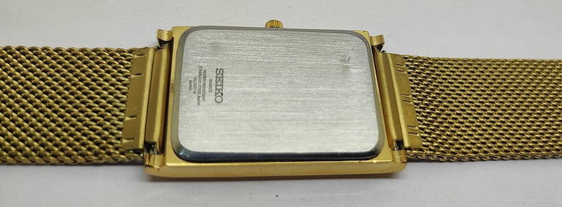 Peut inclure: Une montre Seiko dor&eacute;e avec un cadran rectangulaire et un bracelet en maille. Le bo&icirc;tier de la montre est dor&eacute;, et le dos est argent&eacute; avec le logo et le texte Seiko. Le bracelet en maille est &eacute;galement dor&eacute; et semble &ecirc;tre en m&eacute;tal.