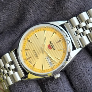 May include: A Seiko 5 automatic wristwatch with a gold-toned dial featuring a textured horizontal line pattern, silver-tone hands and hour markers, and a day-date window at the three o'clock position. The watch has a stainless steel link bracelet.