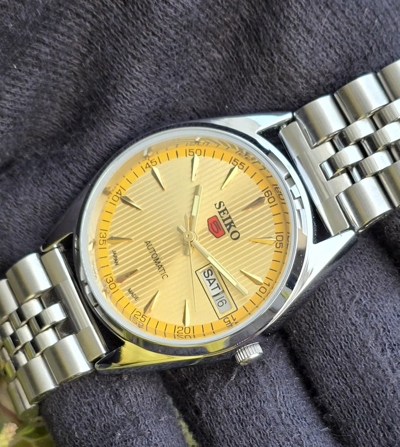 May include: A Seiko 5 automatic wristwatch with a gold-toned dial featuring a textured horizontal line pattern, silver-tone hands and hour markers, and a day-date window at the three o'clock position. The watch has a stainless steel link bracelet.