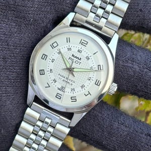 May include: Silver wristwatch with a white face and a silver link strap. The watch face has black numerals, a second hand, and the text "hmt PILOT PARA SHOCK 17 JEWELS INDIA". The watch is shown on a dark surface.