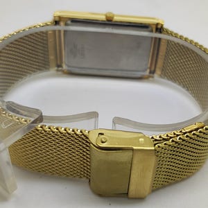 Peut inclure: Une montre-bracelet dor&eacute;e avec un cadran rectangulaire et un bracelet en maille. La montre est dot&eacute;e d'une fermeture &agrave; boucle et est pr&eacute;sent&eacute;e sur un support en plastique transparent. Le bracelet a un motif tiss&eacute;.