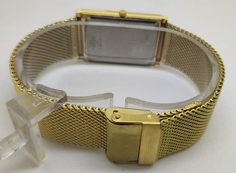 Peut inclure: Une montre-bracelet dor&eacute;e avec un cadran rectangulaire et un bracelet en maille. La montre est dot&eacute;e d'une fermeture &agrave; boucle et est pr&eacute;sent&eacute;e sur un support en plastique transparent. Le bracelet a un motif tiss&eacute;.