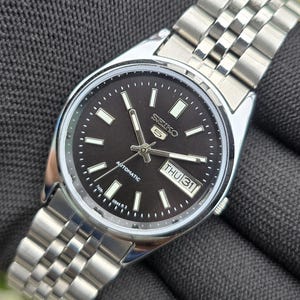 Op de afbeelding: Een Seiko 5 automatisch polshorloge met een ronde zwarte wijzerplaat, zilverkleurige uurmarkeringen en lichtgevende wijzers. Het horloge heeft een dag-datumvenster op de drie-uurpositie met THU 31 en een schakelarmband van gepolijst roestvrij staal.