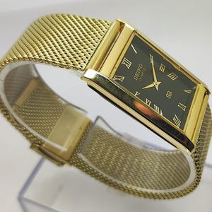 Peut inclure: Une montre Seiko Quartz dor&eacute;e avec un bracelet en maille. Le cadran rectangulaire est vert fonc&eacute; avec des chiffres romains dor&eacute;s. Le bracelet est en maille tiss&eacute;e.