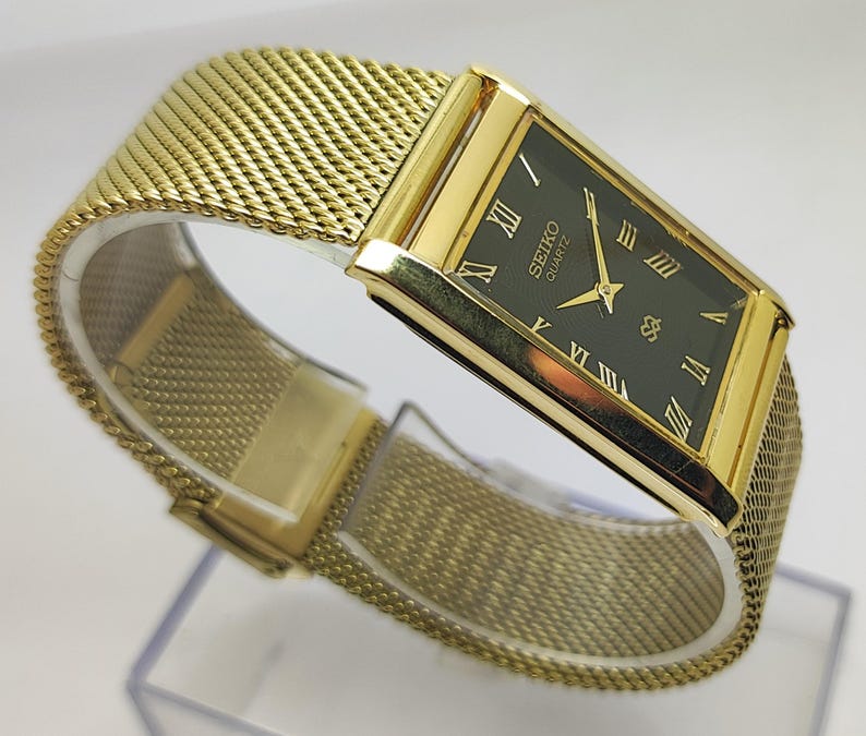 Peut inclure: Une montre Seiko Quartz dor&eacute;e avec un bracelet en maille. Le cadran rectangulaire est vert fonc&eacute; avec des chiffres romains dor&eacute;s. Le bracelet est en maille tiss&eacute;e.