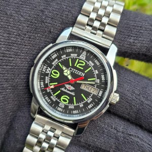 Könnte beinhalten: Eine Citizen Automatik-Armbanduhr mit schwarzem Zifferblatt, leuchtend grünen Ziffern und einem roten Sekundenzeiger. Die Uhr verfügt über ein Edelstahlarmband und eine Datumsanzeige. Die Uhr wird auf einer dunkelgrauen Oberfläche gezeigt.