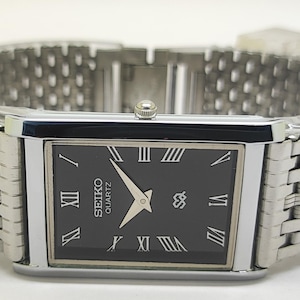 May include: A rectangular Seiko Quartz watch with a black face and silver hands. The watch has Roman numeral hour markers and a silver-toned, woven metal strap. The watch face displays the Seiko logo.