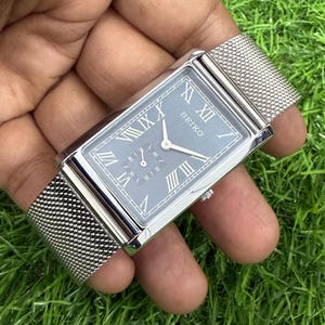 Op de afbeelding: Een rechthoekig Seiko polshorloge met een grijze wijzerplaat en een zilveren mesh band. De wijzerplaat heeft Romeinse cijfers en twee kleinere wijzerplaten. Het horloge ligt op een achtergrond van groen gras.