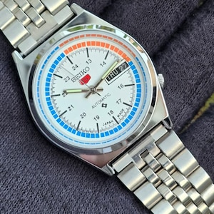 May include: A silver-toned Seiko automatic wristwatch with a white face, featuring blue and orange accents. The watch displays the day and date, with a silver band. The watch face includes the text "SEIKO" and "AUTOMATIC".
