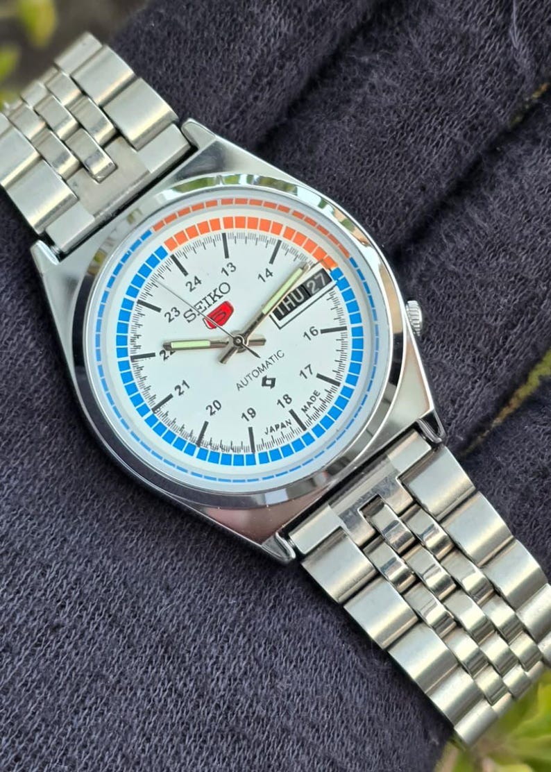 May include: A silver-toned Seiko automatic wristwatch with a white face, featuring blue and orange accents. The watch displays the day and date, with a silver band. The watch face includes the text "SEIKO" and "AUTOMATIC".