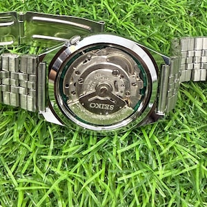 May include: A silver-toned Seiko wristwatch with a clear back, showcasing the intricate internal mechanism. The watch has a silver band and is set against a backdrop of green grass.