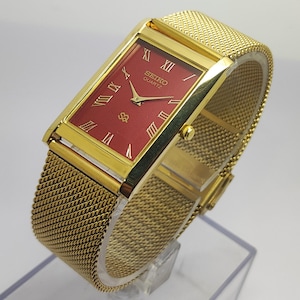 Puede incluir: Un reloj Seiko Quartz dorado con una esfera rectangular roja y números romanos dorados. El reloj tiene una correa de malla y se muestra sobre un soporte transparente. La esfera del reloj tiene el logotipo de Seiko Quartz.