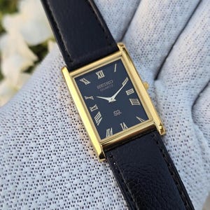 Könnte beinhalten: Eine rechteckige, goldfarbene Seiko Quartz-Uhr mit schwarzem Zifferblatt und schwarzem Lederarmband. Das Zifferblatt der Uhr hat goldene römische Ziffern und Zeiger. Die Uhr wird auf einer weißen Oberfläche präsentiert.