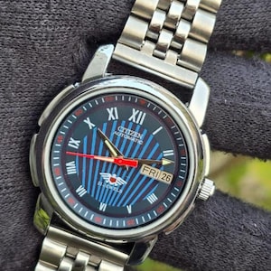 May include: A Citizen Automatic wristwatch with a silver-tone stainless steel band. The watch face is black with blue and red accents, including the day and date. The watch displays the day as "FRI 26".