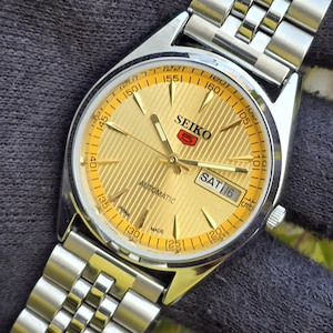 May include: A Seiko 5 automatic wristwatch with a gold-toned dial featuring a textured striped pattern, silver-tone hands and hour markers, and a day-date display at the three o'clock position. The watch includes a stainless steel link bracelet.