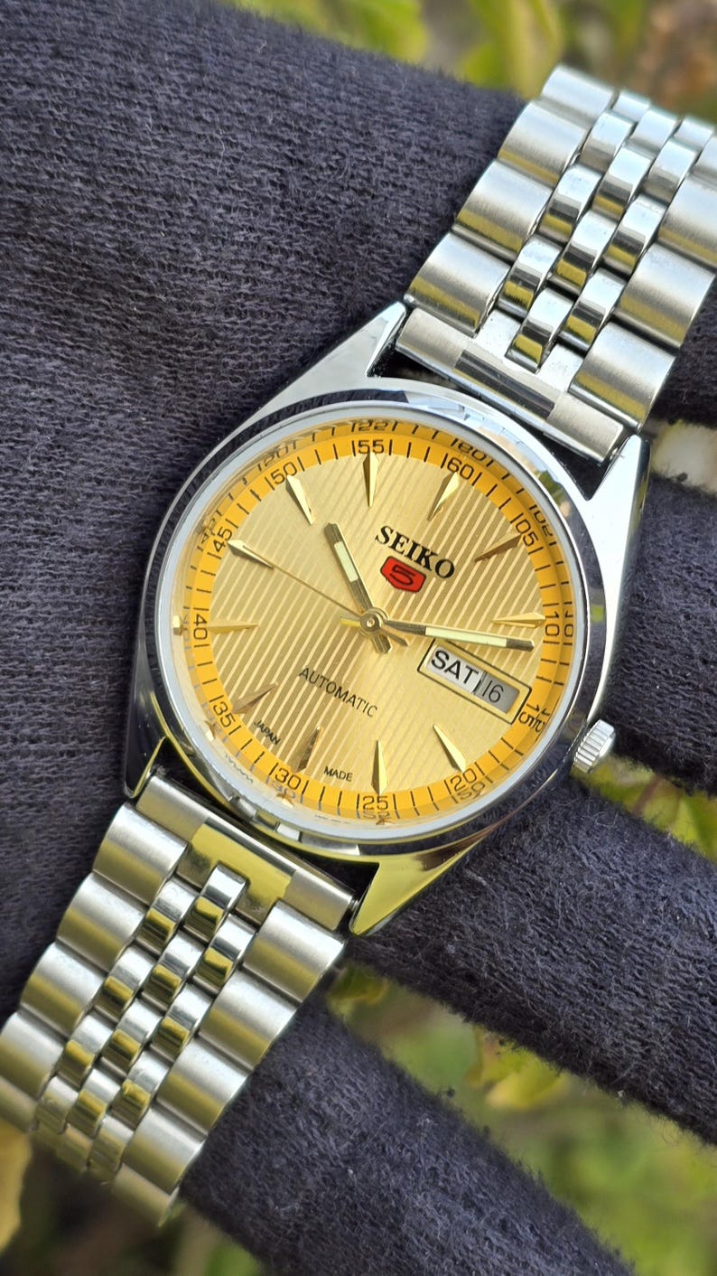May include: A Seiko 5 automatic wristwatch with a gold-toned dial featuring a textured striped pattern, silver-tone hands and hour markers, and a day-date display at the three o'clock position. The watch includes a stainless steel link bracelet.
