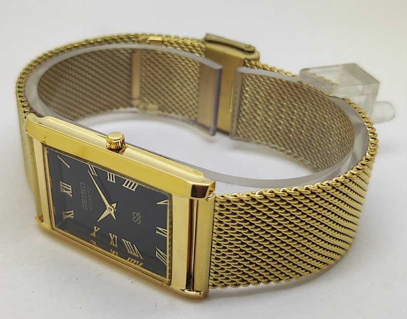 Peut inclure: Une montre Seiko Quartz dor&eacute;e avec un cadran noir rectangulaire et des chiffres romains dor&eacute;s. La montre est dot&eacute;e d'un bracelet en maille dor&eacute;e. Le cadran affiche le logo Seiko et le mot "Quartz".