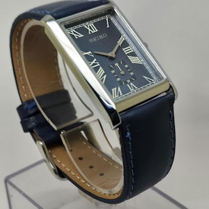 May include: A rectangular Seiko watch with a navy blue face and silver casing. The watch features Roman numeral hour markers and a small seconds dial. The navy blue leather band is stitched with blue thread.