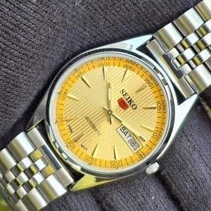 May include: A Seiko 5 automatic wristwatch featuring a gold-toned dial with a textured horizontal line pattern, silver-toned hands and hour markers, and a day-date display window. The watch has a stainless steel case and a multi-link metal bracelet.