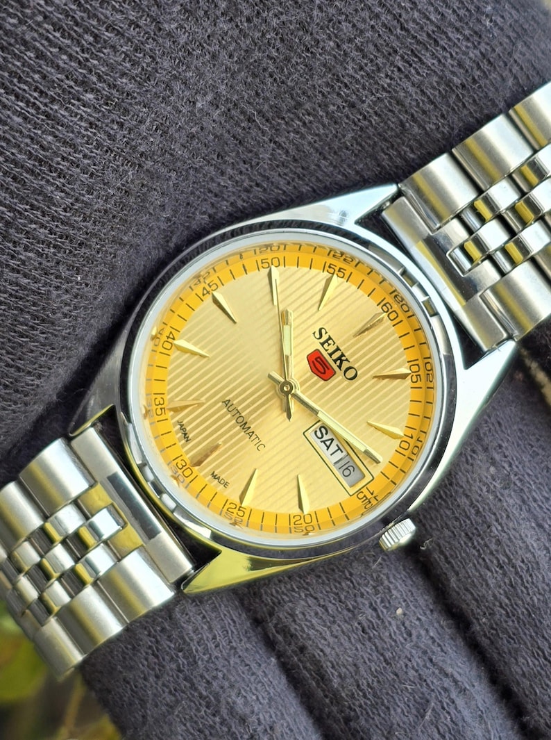 May include: A Seiko 5 automatic wristwatch featuring a gold-toned dial with a textured horizontal line pattern, silver-toned hands and hour markers, and a day-date display window. The watch has a stainless steel case and a multi-link metal bracelet.