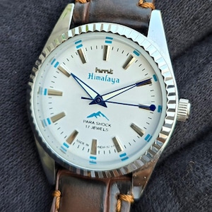 May include: A vintage HMT Himalaya wristwatch with a white face, blue detailing, and a brown leather strap. The watch face displays the text "Himalaya" and "Para Shock 17 Jewels". The watch has a silver bezel and case.
