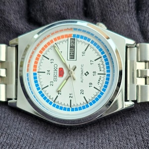 May include: A silver-toned Seiko automatic wristwatch with a white face. The watch features a day-date display, a blue and orange accent ring, and a stainless steel band. The watch face displays the Seiko logo and the words "AUTOMATIC" and "MADE IN JAPAN".
