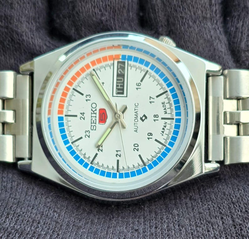 May include: A silver-toned Seiko automatic wristwatch with a white face. The watch features a day-date display, a blue and orange accent ring, and a stainless steel band. The watch face displays the Seiko logo and the words "AUTOMATIC" and "MADE IN JAPAN".