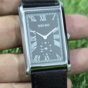 Peut inclure: Une montre Seiko rectangulaire avec un cadran noir et un boîtier argenté. La montre présente des chiffres romains pour les heures et un bracelet en cuir noir. La montre est tenue sur un fond d'herbe verte.
