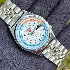 May include: A silver-toned Seiko automatic wristwatch with a white face, featuring blue and orange accents. The watch displays the day and date, with a silver metal band. The watch face includes the text "SEIKO AUTOMATIC MADE IN JAPAN".