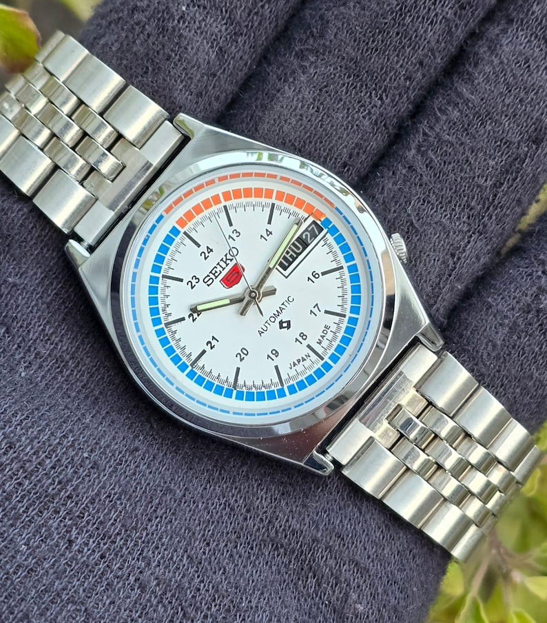 May include: A silver-toned Seiko automatic wristwatch with a white face, featuring blue and orange accents. The watch displays the day and date, with a silver metal band. The watch face includes the text "SEIKO AUTOMATIC MADE IN JAPAN".
